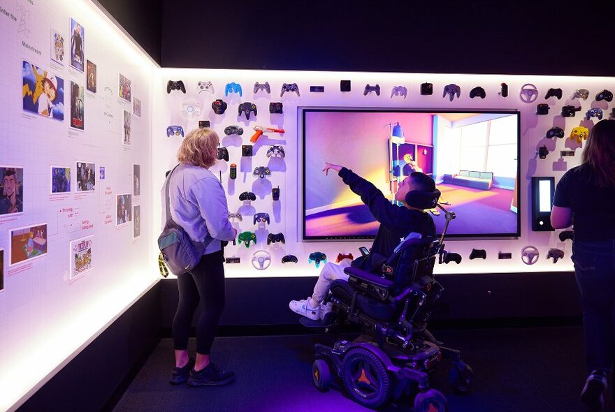 A person in a wheelchair pointing at a gallery display of video game controllers.