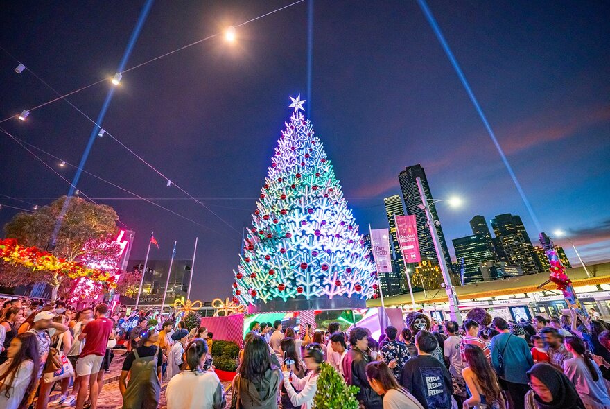 Crowds milling around a colourful LED Christmas tree at night.