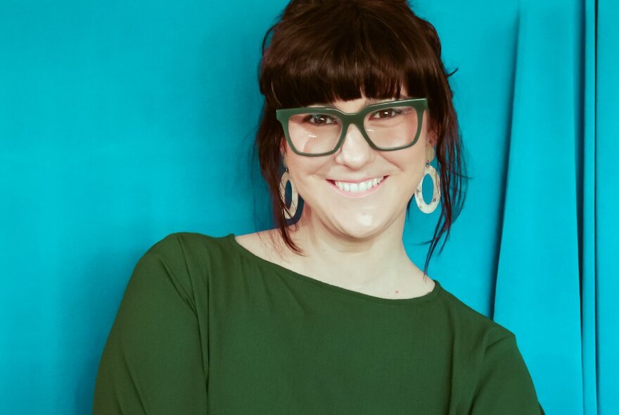 A portrait of comedian, Rebecca Perich, wearing a green top and glasses, in front of a bright blue background.