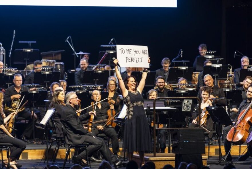 An orchestra on a stage with their instruments, with a singer in front of them holding up a sign that reads TO ME YOU ARE PERFECT.