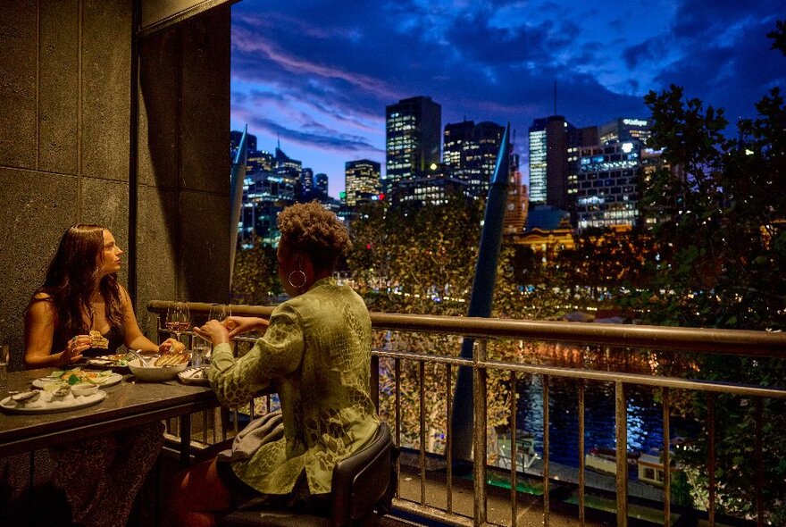 Two friends are eating and drinking on a terrace bar. There is a city skyline in the background. 