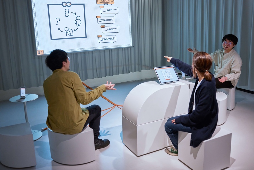 Three people sitting on white cuboid stools in a sterile-looking room with a projector screen and curtains.