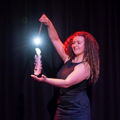 A magician on stage pointing a light-up wand at a large feather standing upright in her hand.