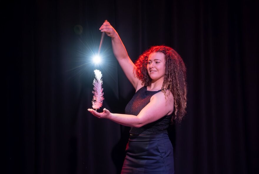 A magician on stage pointing a light-up wand at a large feather standing upright in her hand.