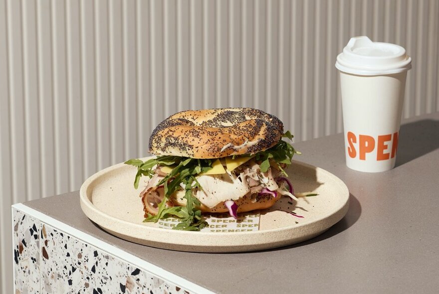 A filled bagel on a plate next to a takeaway coffee.