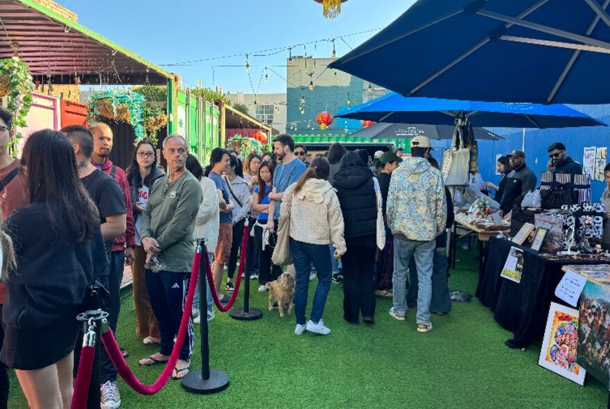 People queuing on astroturf next to market stalls with big umbrellas.