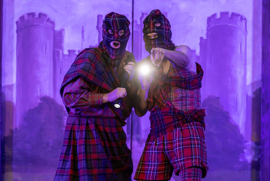 Two tartan-clad performers with balaclavas and a torch, one of them pointing, in front of a projected image of a castle. 