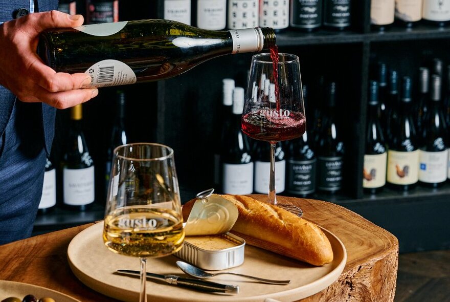 A waiter pouring red wine into a glass next to a plate with a baguette and an open tin of pate.