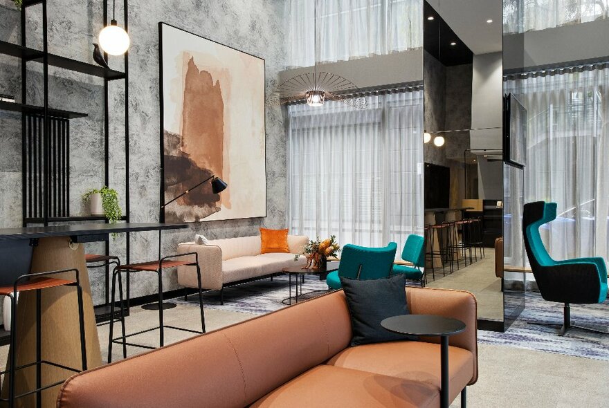 Hotel foyer with grey walls with black shelves and brown and blue seating, large painting on the wall and windows draped in white curtains.