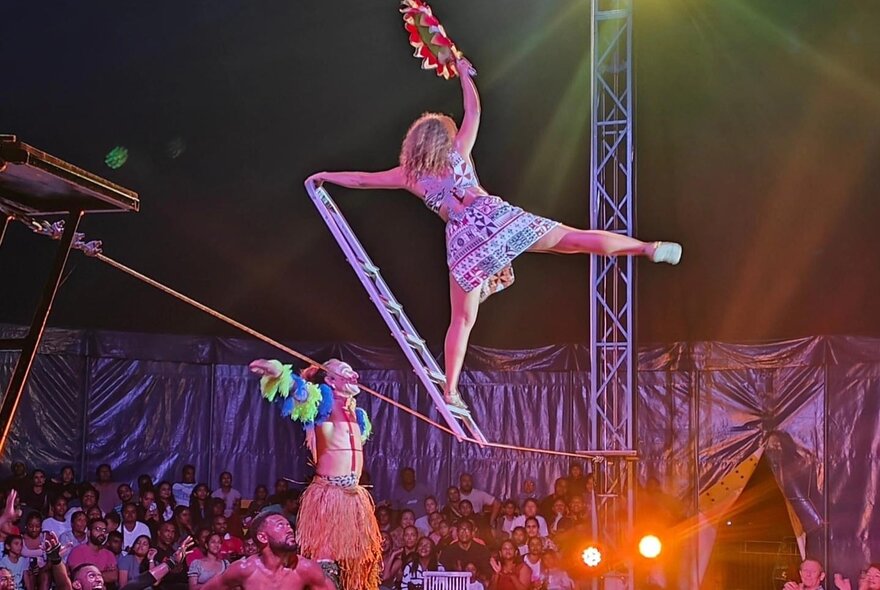 A circus performer on a ladder balanced on a wire in front of an audience. 
