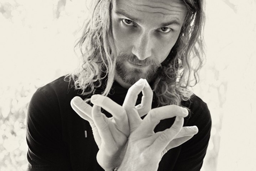 Black and white portrait of a man with long hair and both hands intertwined, making shapes with his oddly bent fingers.