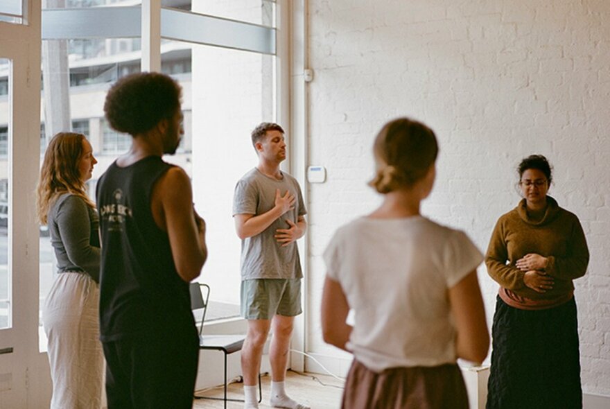 People standing quietly in a well-lit room, their eyes closed, hands on their chests and bellies.