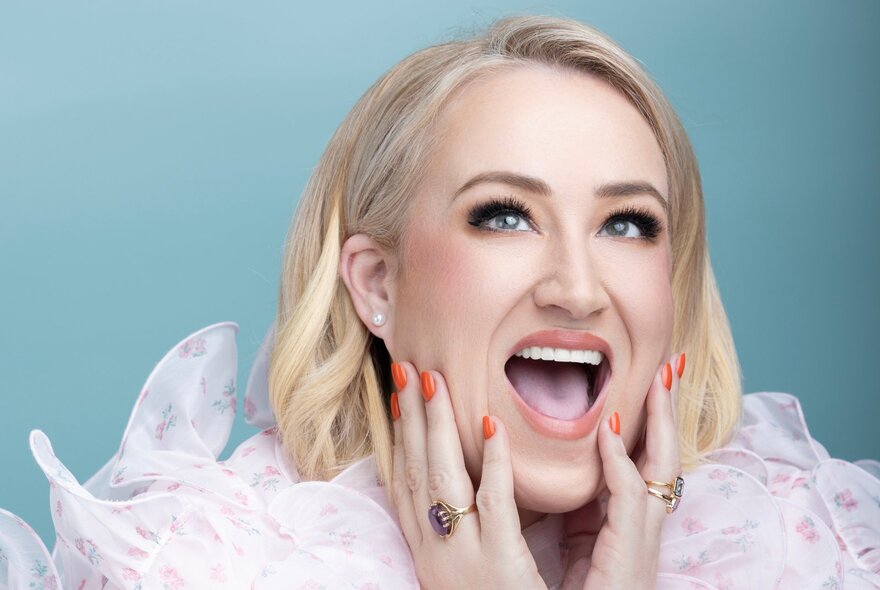 Comedian Rosie Waterland, with her hands resting on her jaw, her mouth is open as if laughing, she has bob-length blond hair and billowing fabric around her neck.