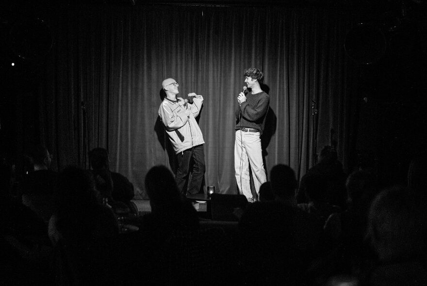 Two stand-up comedians on a stage facing each other, each talking into microphones, in front of an audience. 