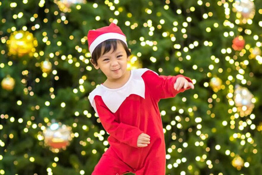 Smiling pre-school aged child dressed in a red and white onesie and wearing a red Santa style hat on their head, dancing in front of a large Christmas tree.