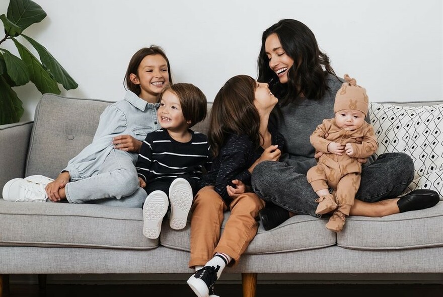 Family group of mother with four children including a young baby, seated on a grey couch.