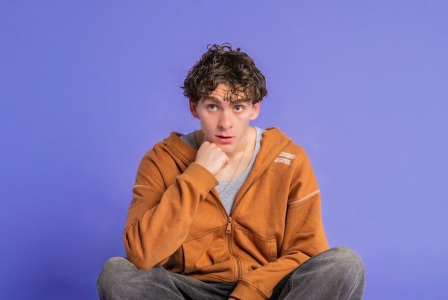 Comedian Louis Prandolini wearing an orange jacket, his hand under his chin with a slightly bemused expression on his face, posed against a blue background.
