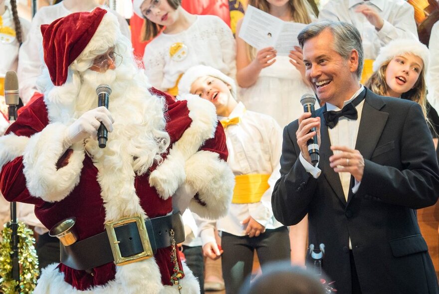 Santa and a man in a black suit, both holding microphones, with a crowd of children behind them, at a Christmas Carol concert.