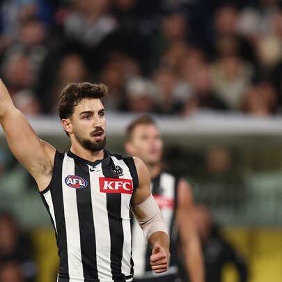 A Collingwood AFL player celebrating on the field with his arm raised high above his head, a blurred stadium crowd behind him. 