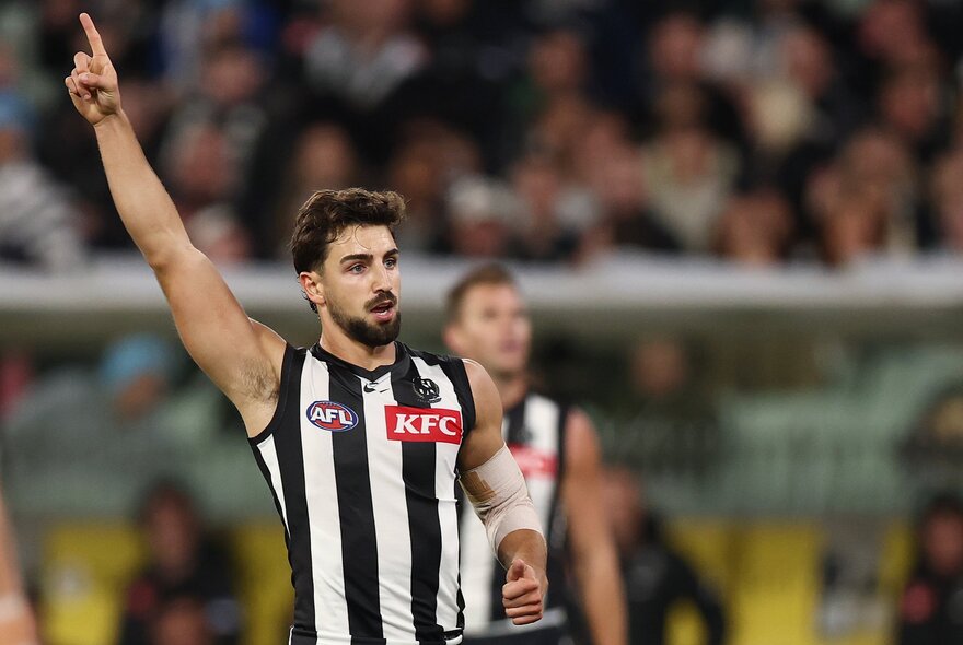 A Collingwood AFL player celebrating on the field with his arm raised high above his head, a blurred stadium crowd behind him. 