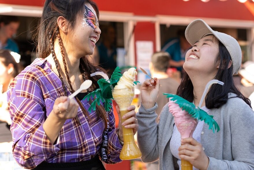 Two friends laughing and holding giant ice-creams in their hands, one of them with a butterfly design painted on her face; crowds in the background.