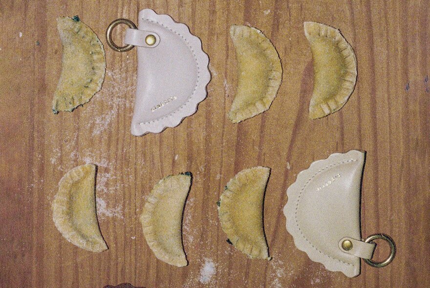 Looking down at two rows of half-moon shaped agnolotti on a wooden kitchen board, with two small pale leather coin purses of the same shape also lying on the board.