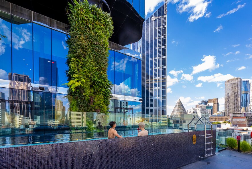 Two friends are sitting in a rooftop pool with a view of Melbourne's skyline