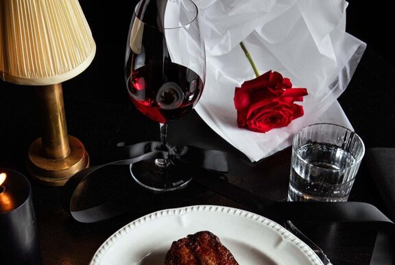 A red rose, a steak on a white plate, a glass of red wine and a small table lamp on a restaurant table.