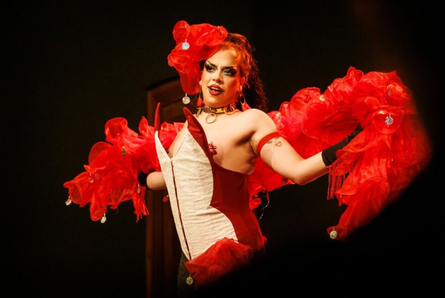 Drag cabaret performer on a dark stage wearing a red feather stole and hat, bustier and heavy makeup.