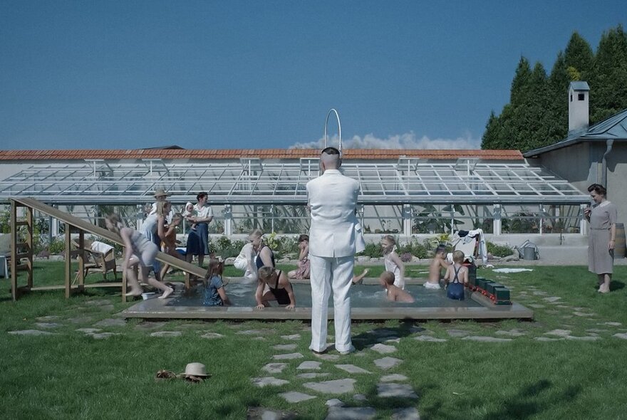 A still from the move Zone of Interest with children playing in a pool in a large garden, there is a glasshouse behind them and a man wearing a white suit with his back to the viewer is facing the pool.