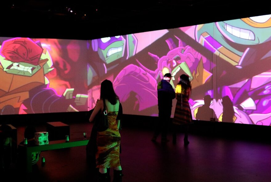 People walking around a darkened exhibition space with images from the TV show Teenage Mutant Ninja Turtles projected onto the walls around them.