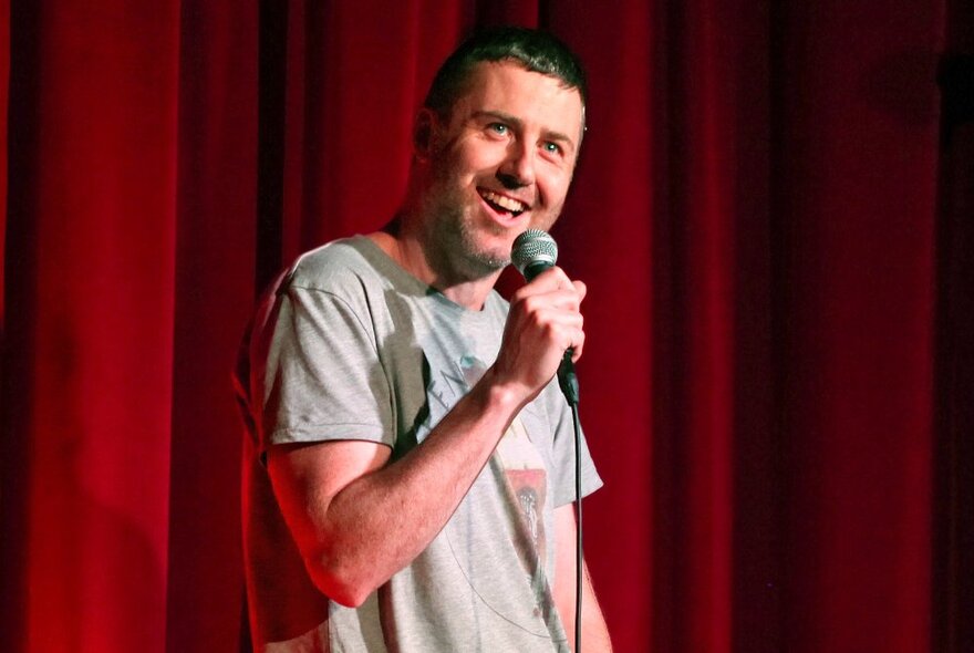 Comedian Lloyd Langford holding a microphone in his hand and performing stand-up comedy on a stage against a red curtain.