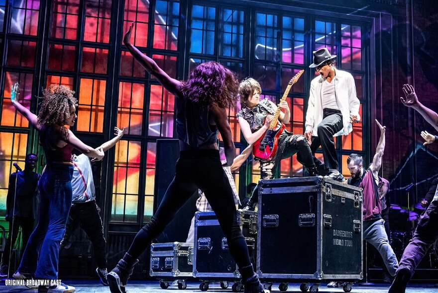 A dancer on stage in a white jacket and black pants, in the role of Michael Jackson, with musicians and dancers alongside, in the stage production of MJ the Musical.