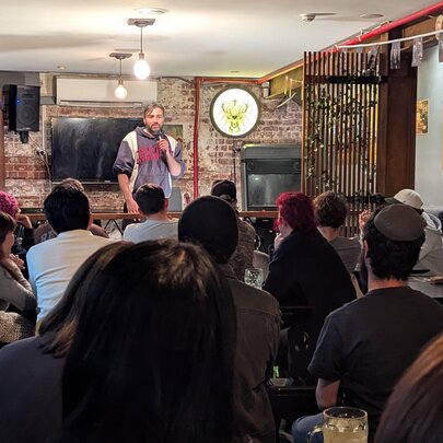 Seated audience watching a standup comedian perform with microphone in a small underground venue.