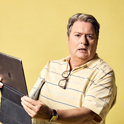 Comedian Dave O'Neil holding na laptop with spectacles hanging from his striped polo shirt, looking aghast.