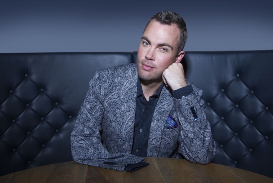 Pianist Nicholas McCarthy seated in a leather booth with his head leaning on his hand, looking relaxed in a paisley jacket.