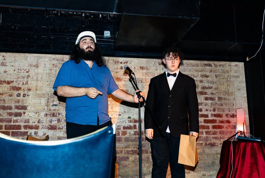 Two people on a stage in front of a fake brick wall and microphone, one in a tuxedo. 