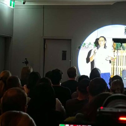 Comedian performing on a small stage, spotlit in front of seated audience.