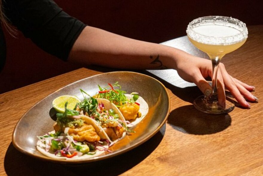 Three tacos on a plate with a hand holding a salt-rimmed margarita