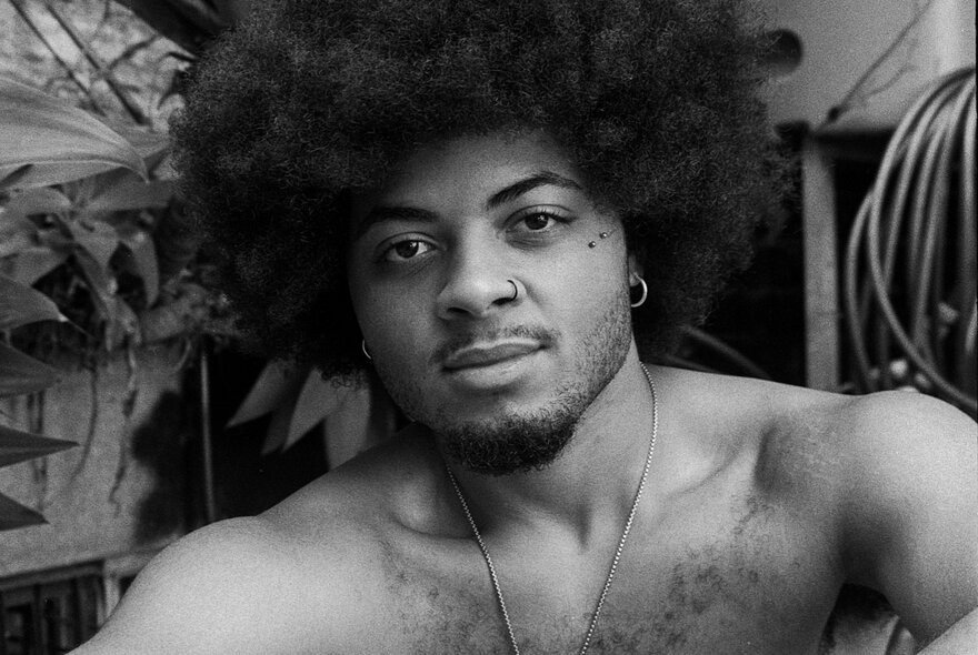 A black-and-white portrait of a shirtless person with a large afro, a nose ring, a goatee, and a thin necklace.