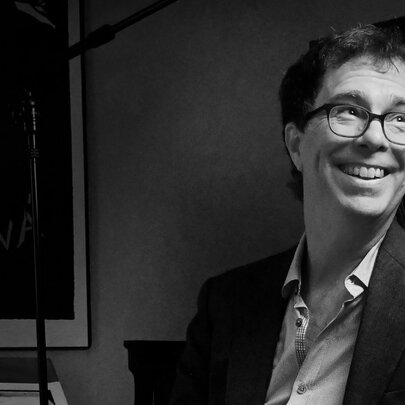 Black and white half portrait of American singer, songwriter, and pianist Ben Folds, a middle-aged man wearing an open shirt and suit jacket. 