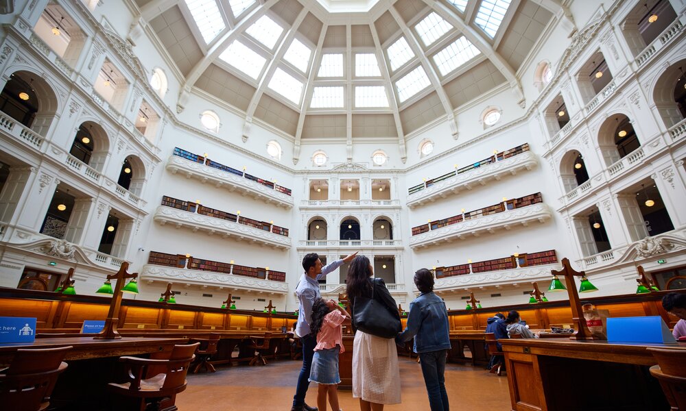 A family of four standing in a grand library with antique desks, large white walls and a dome ceiling.