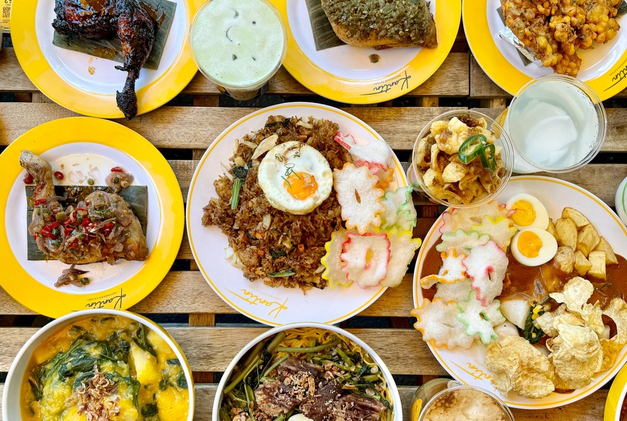Overhead view of a wooden table filled with various colourful plates and bowls of traditional Indonesian food, including fried rice with an egg, chicken dishes, and side dishes.