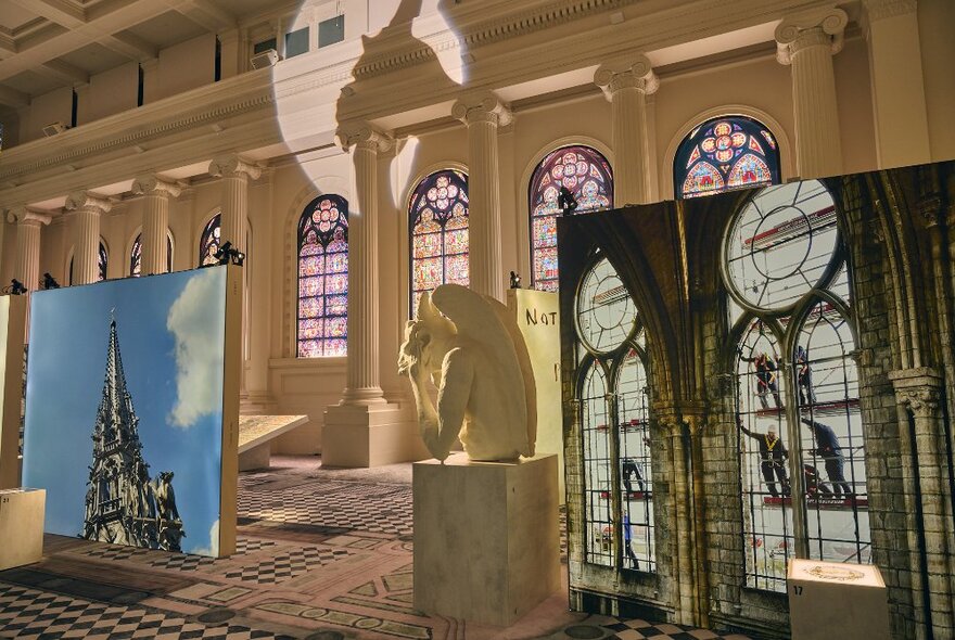 Large scale prints showing parts of La Notre-Dame de Paris, inside a room with high ceilings and a row of stained glass arched windows behind.