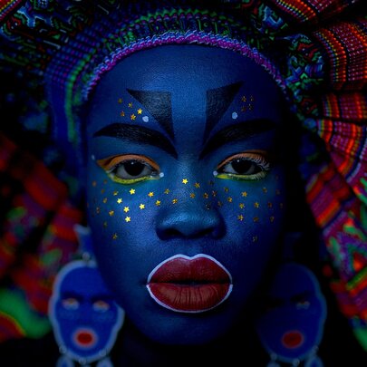 Portrait of a woman with her face painted blue and embellished with gold stars, wearing a colourful, fan-like headdress.