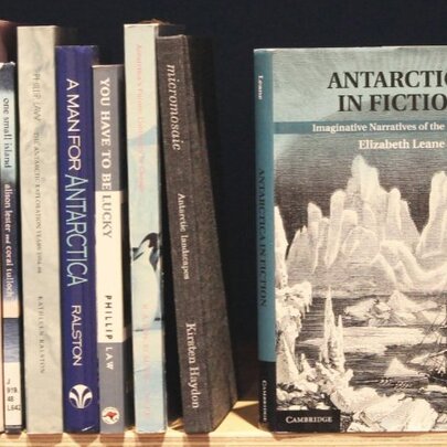 A selection of books on a shelf, spine out, with one of the books called 'Antarctica In Fiction' with its front cover facing the viewer. 
