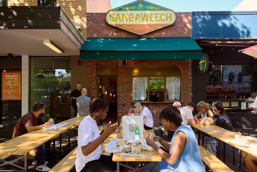 People eating on benches outside Sangaweech in dappled light. 