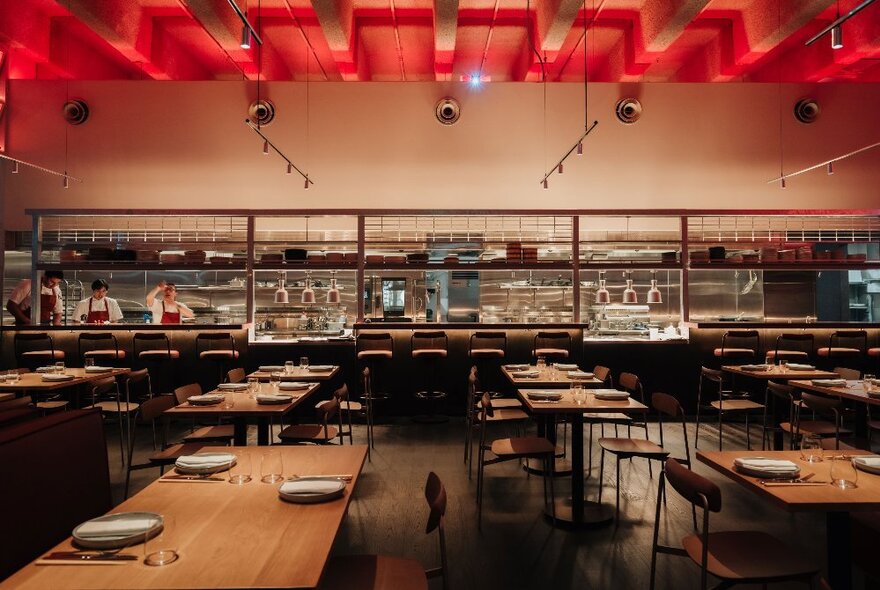 Casual restaurant with long wooden tables and metal chairs, red illuminated ceiling feature and long open kitchen.