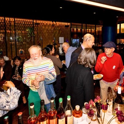 A crowded indoor social gathering featuring people mingling around a bar stocked with wine bottles and plates of food.
