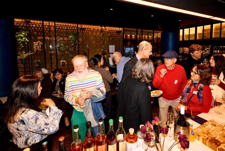 A crowded indoor social gathering featuring people mingling around a bar stocked with wine bottles and plates of food.
